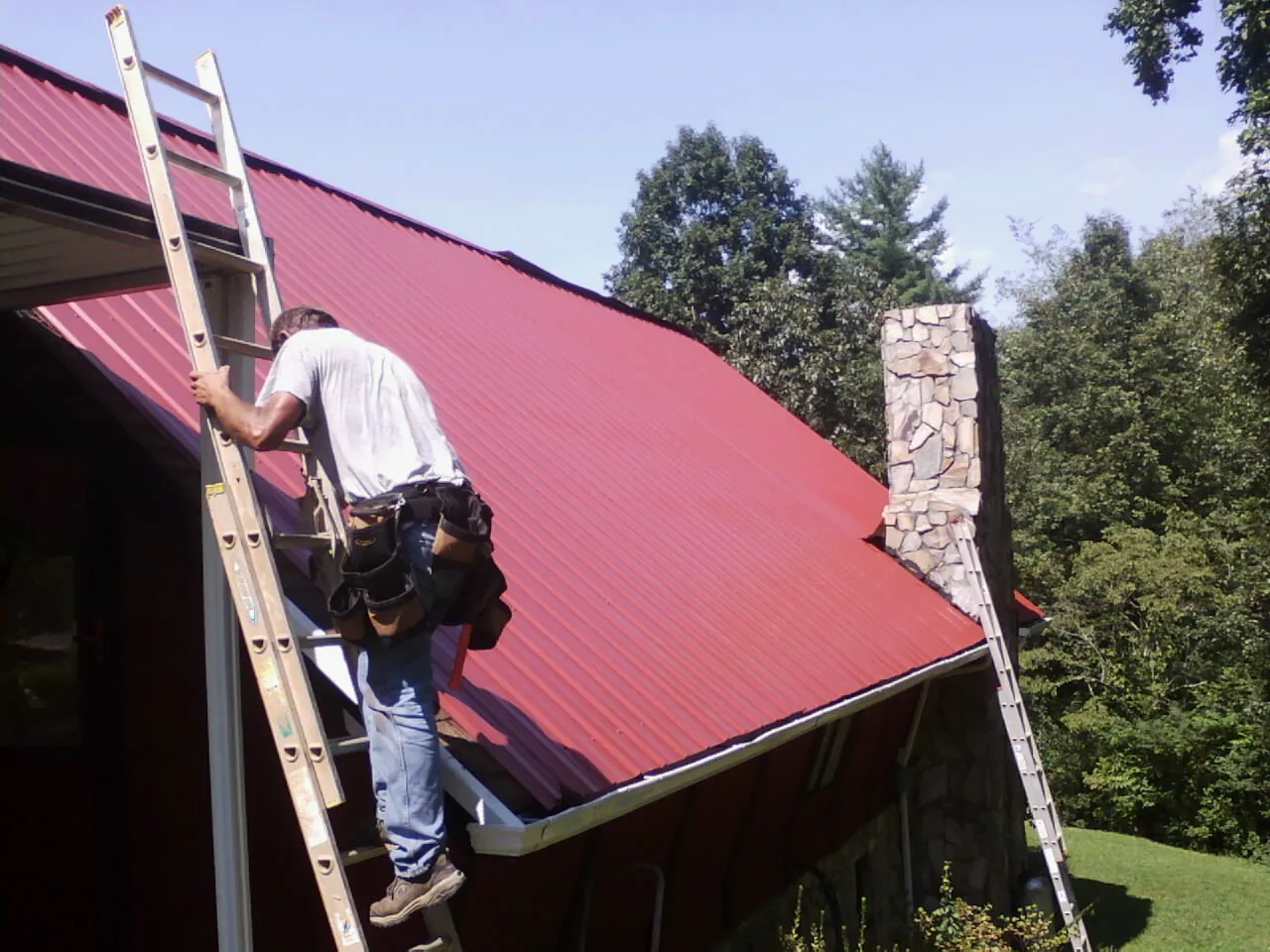 Roofer on a ladder beside a red metal roof and stone chimney — Soffit & Fascia Repair by Roofing Brothers in Wernersville